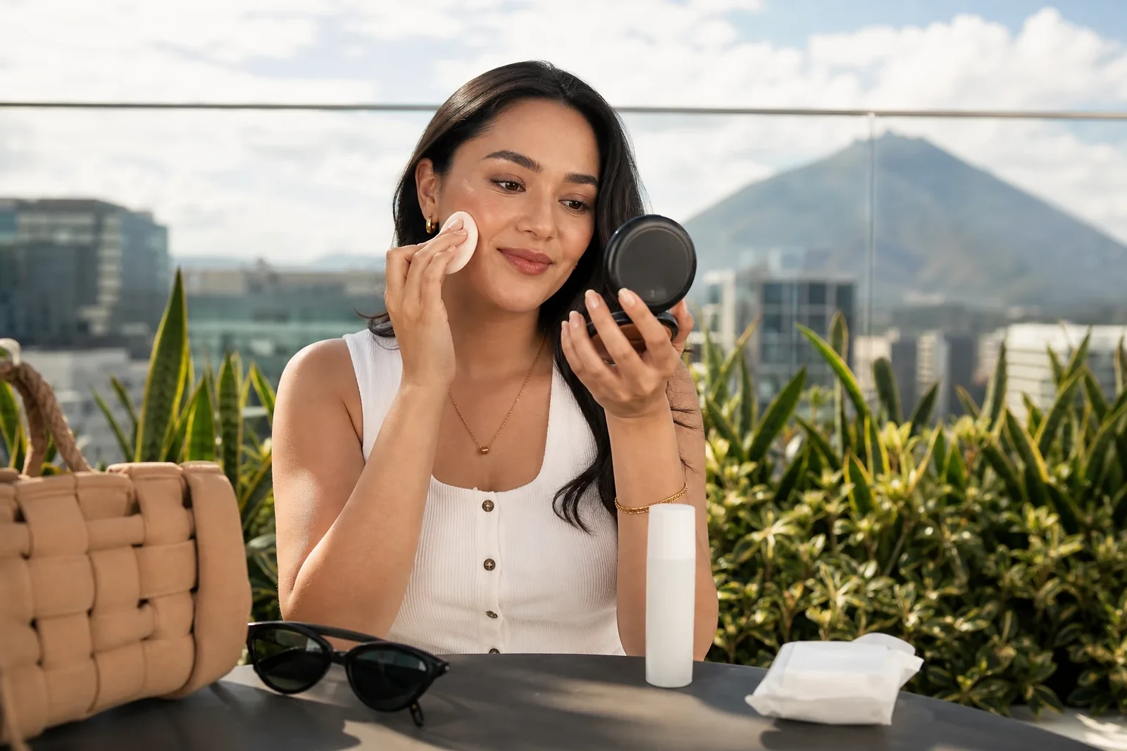 Reaplicar protector solar sobre maquillaje con esponja en la mejilla, mientras una mujer sostiene un compacto en una terraza soleada de Guatemala.