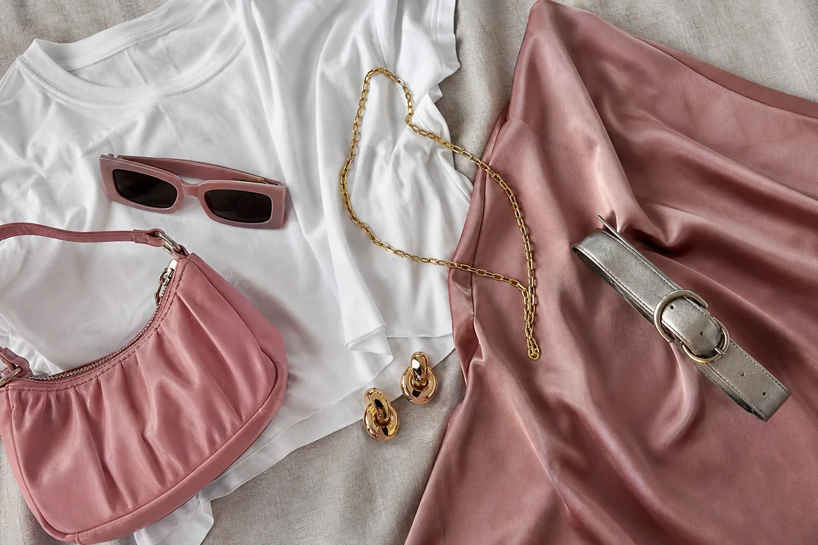 Flat lay de bolso rosa, lentes de color, cinturón metálico, joyería dorada y falda satinada, estilo personal de ZOE Salon Guatemala